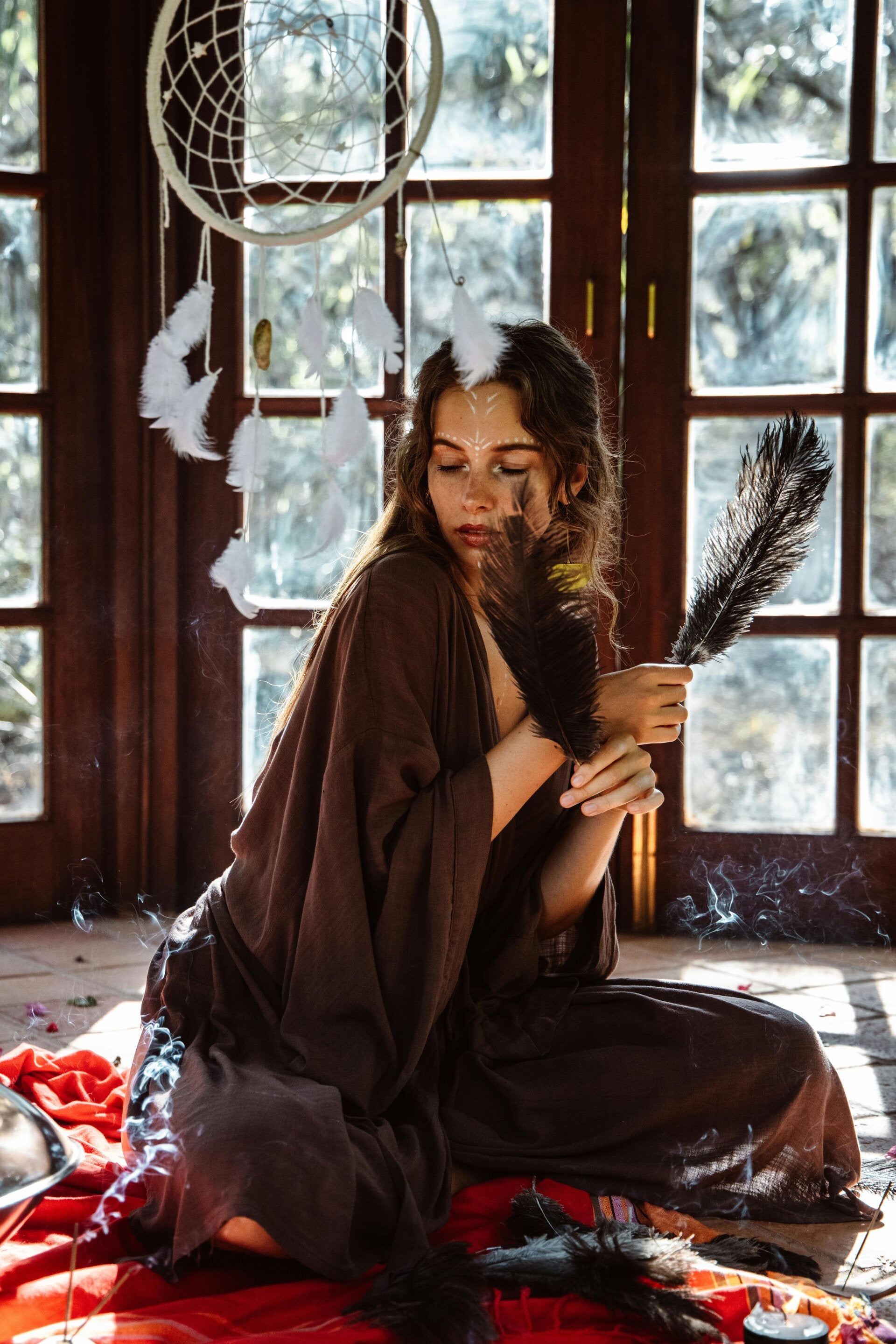 Woman sitting gracefully on the floor, adorned with feathers and surrounded by a dreamcatcher—radiating divine feminine energy and spiritual power.