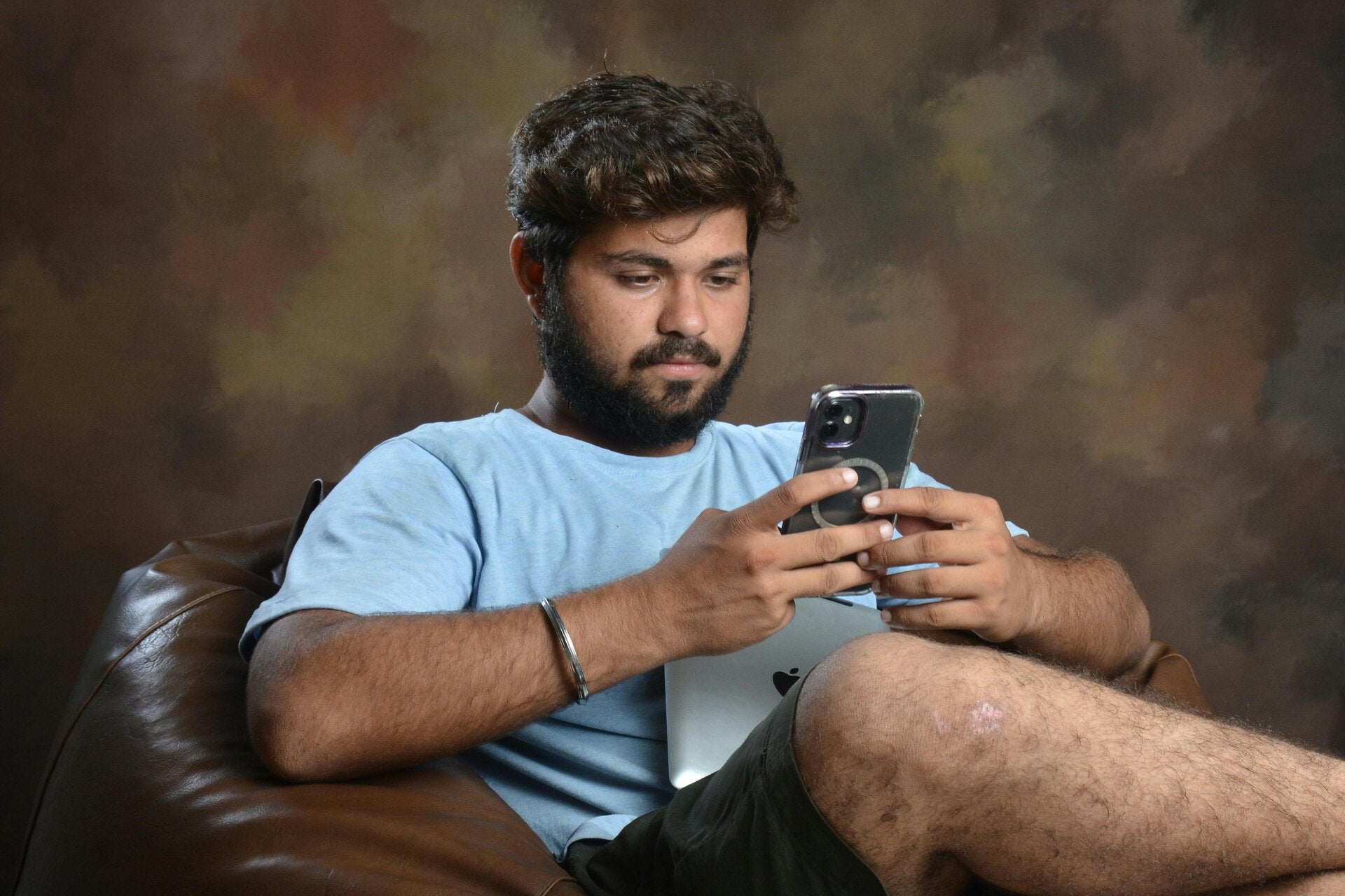 a man in  a light bliue t shirt sits cross-legged in a brown lleather armchair whilst he looks at his mobile phone