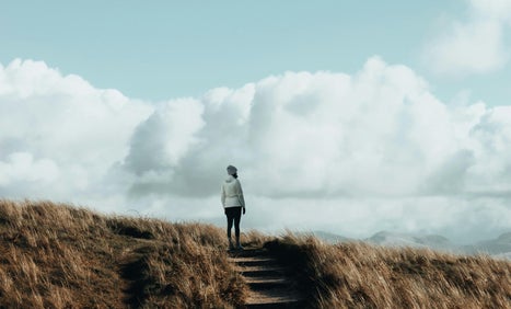 A women in casual warm clothing looking out over a view of hills in the distance after reaching the top of a gentle sloping hill