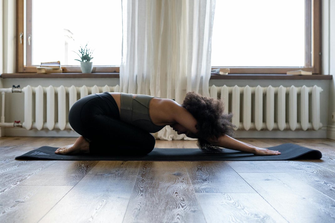 Eine Frau in bequemer Sportkleidung sitzt in der Kindsposition auf einer Yogamatte. Ihr Oberkörper liegt entspannt auf den Oberschenkeln, die Arme sind nach vorne ausgestreckt, Stirn auf der Matte. Die Haltung wirkt beruhigend und dehnt sanft den Rücken