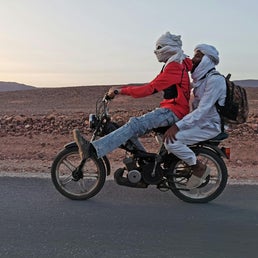Two Moroccan men riding a motorcycle through the desert, wearing traditional Sahara headscarves, showcasing authentic Moroccan culture and nomadic desert lifestyle.