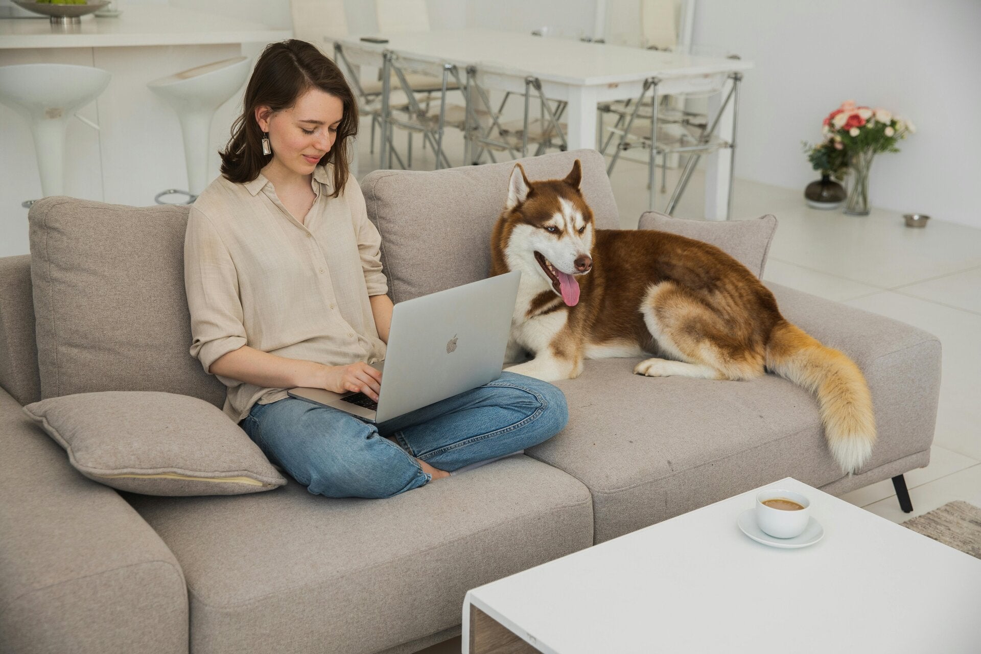 Woman sitting on her couch at home, working on her laptop with her dog beside her, symbolizing comfort, flexibility, and the joy of working from home with pets.