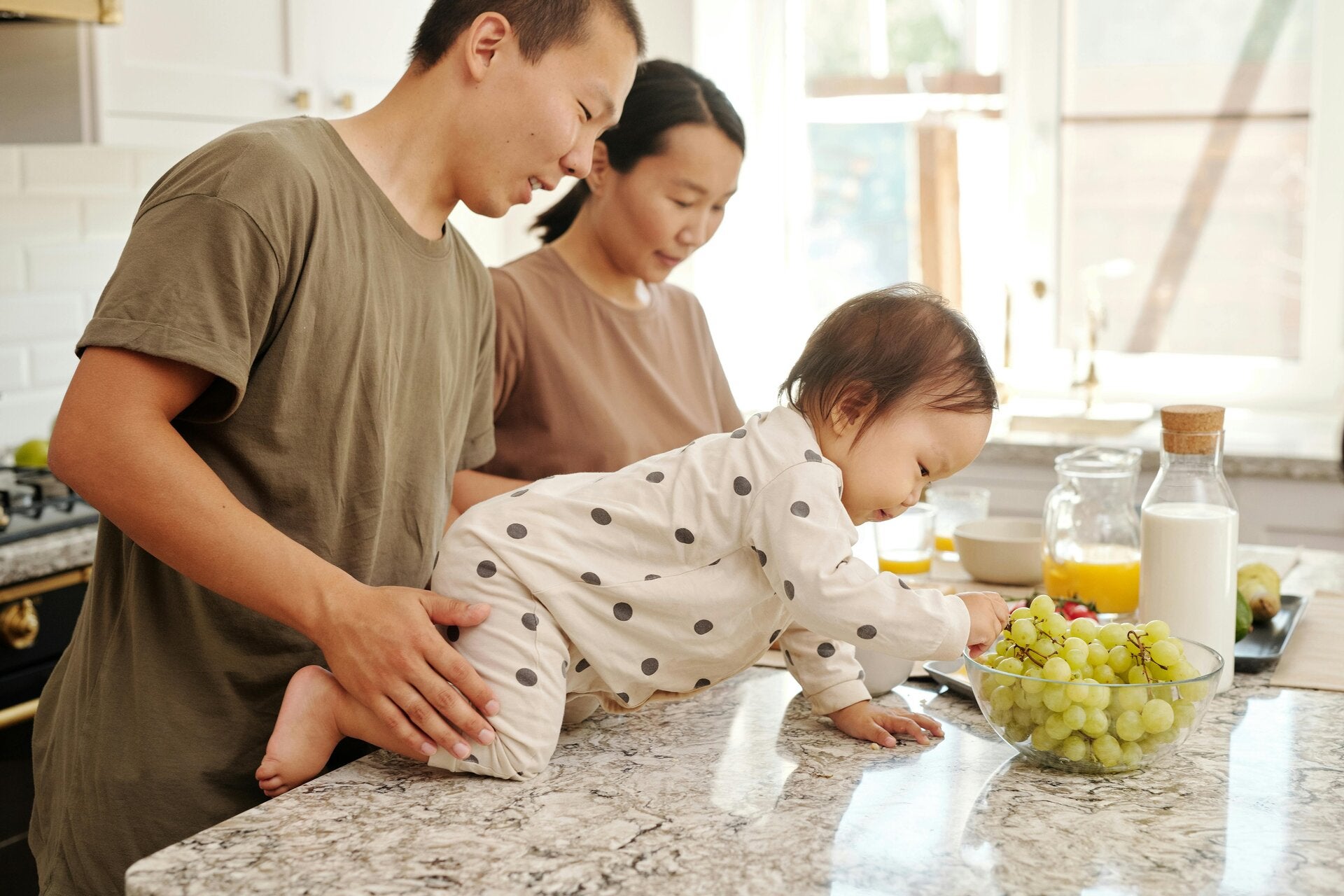 A smiling family gathered in the kitchen with their baby sitting safely on the counter, happily eating a snack, capturing a warm everyday moment of love and togetherness.