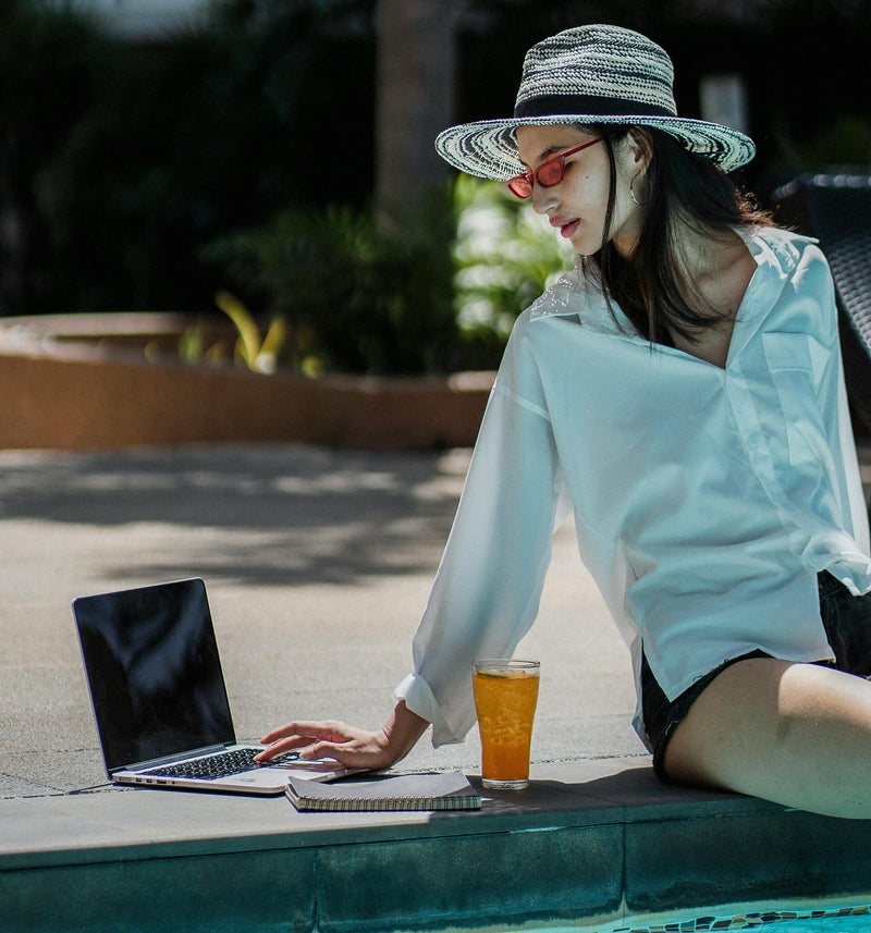 Person wearing sunglasses and a wide-brimmed hat, sitting by a pool with a laptop, notebook, and drink.