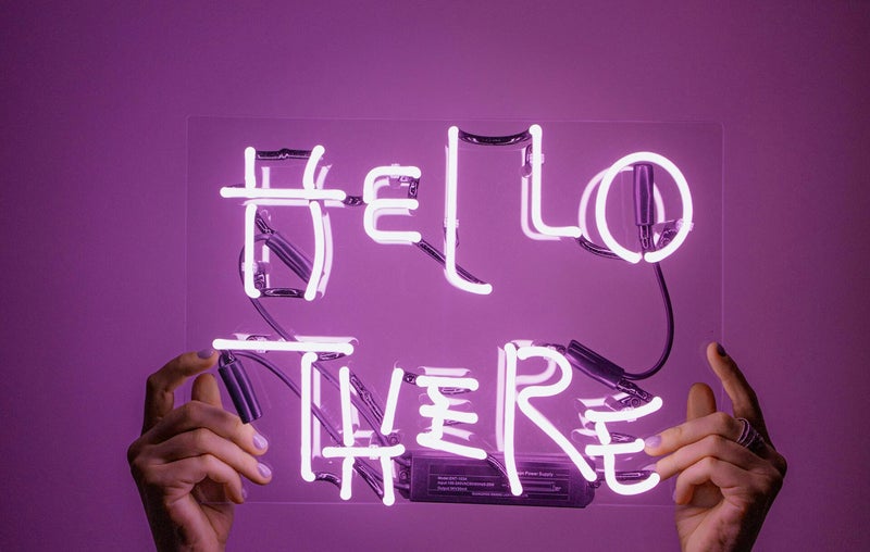 Purple wall with 2 hands holding up a pink neon sign that says ' Hello There'. This demonstracte a welcome to our blog page