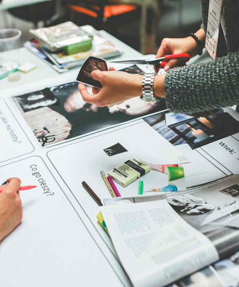 Personen arbeiten gemeinsam an einem Kreativprojekt im Rahmen eines Workshops in einem Büro oder Coworking Space. Auf einem Tisch liegen Magazine, Stifte, Farbmuster und Ausdrucke mit Fotos. Eine Person schneidet ein Bild mit einer Schere aus.