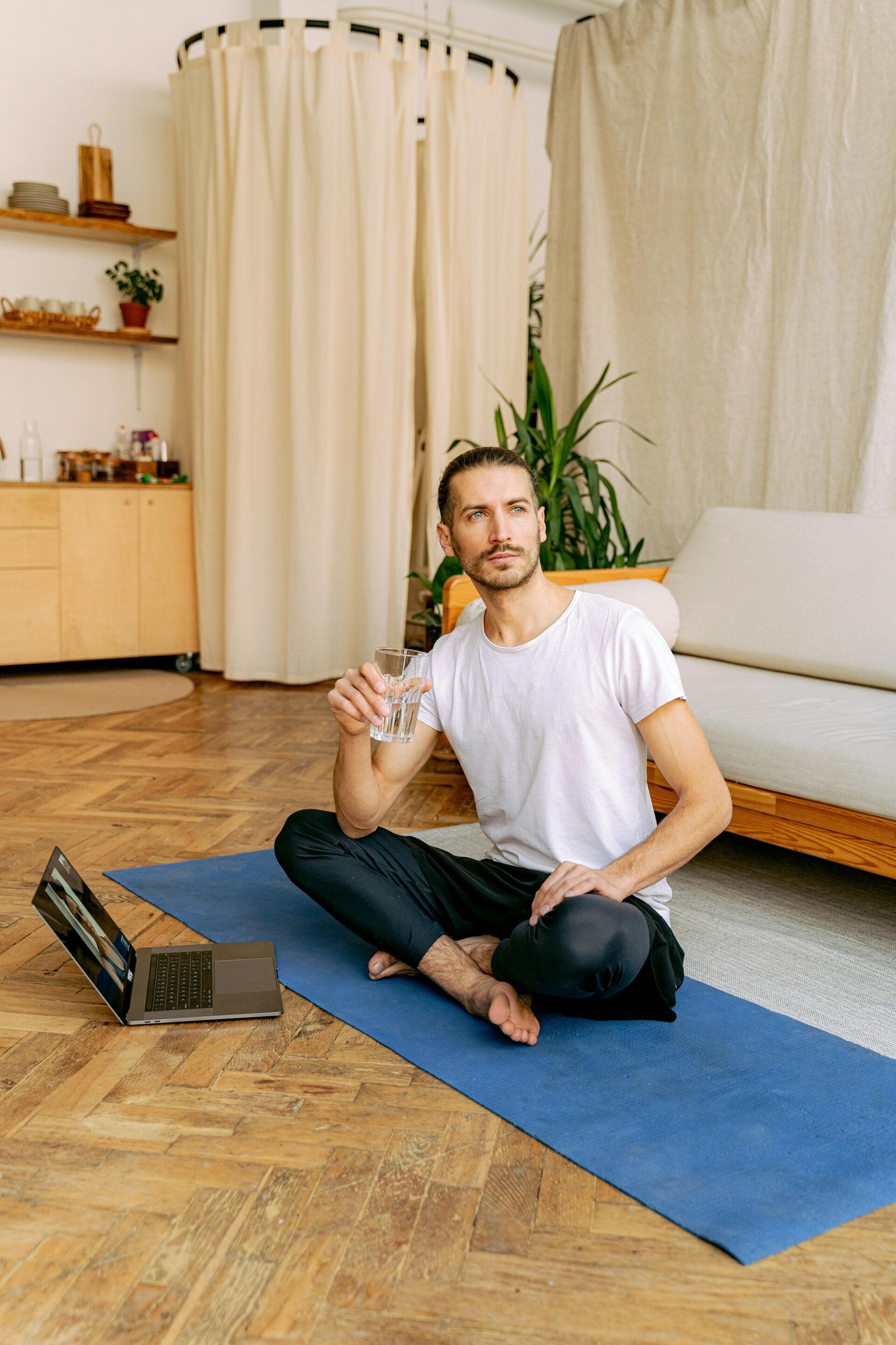 A man sitting comfortably on a yoga mat in a brightly lit room, looking at a laptop screen during an online yoga and coaching session.