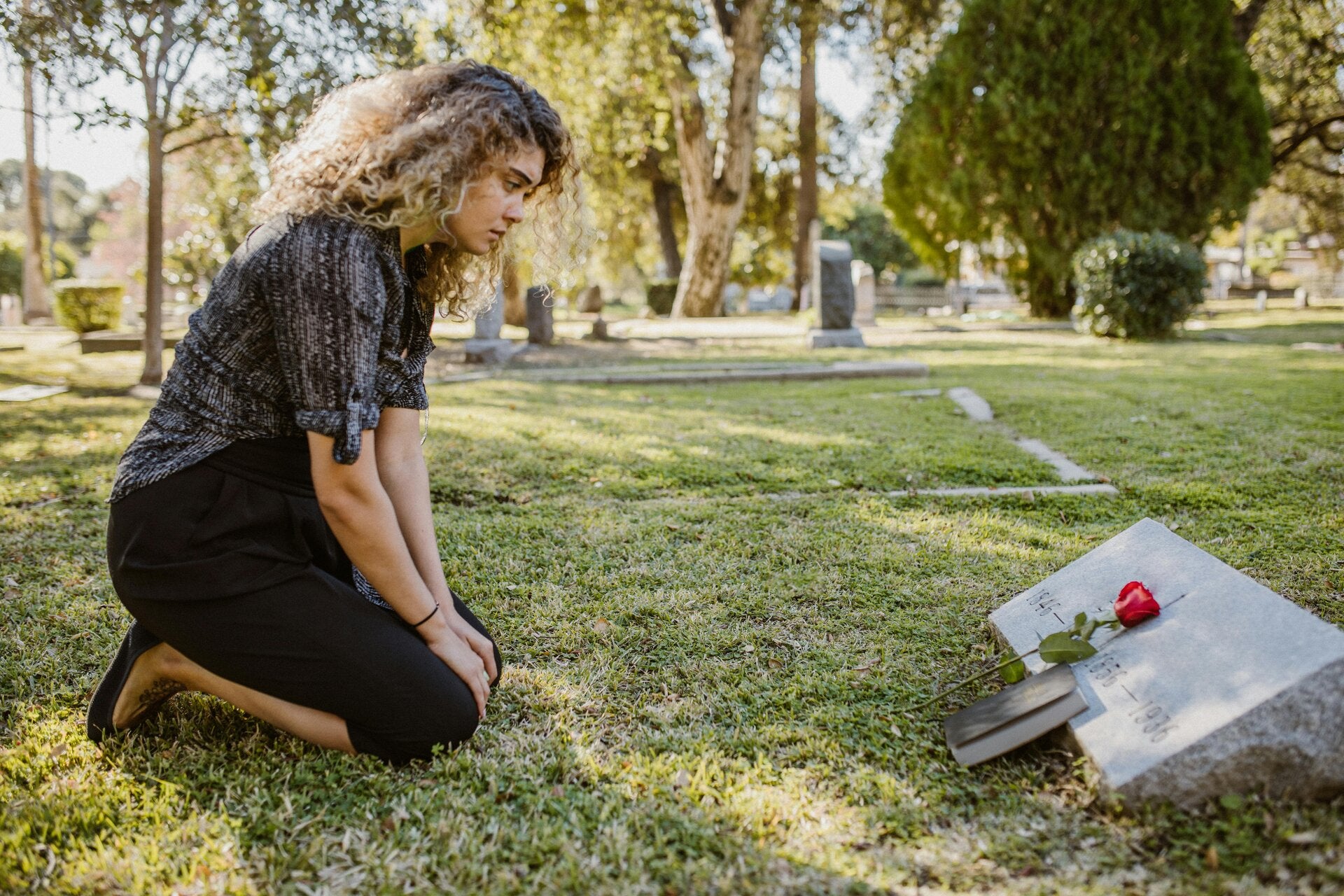 Lady kneeling down facing a grave with a red rose placed on this.