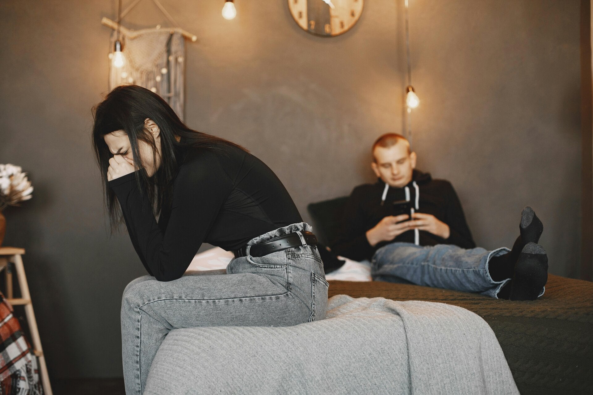 woman sitting at the foot of a bed crying and her partner sitting on his cell phone ignoring her