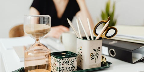 A workspace setting featuring an hourglass, a cup filled with writing utensils, and a person seated at the desk, creating a focused and contemplative atmosphere