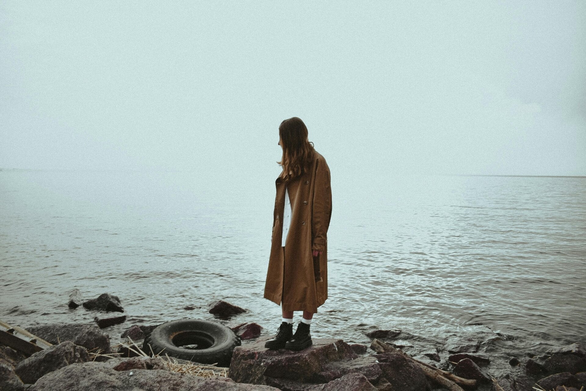 Eine einzelne Frau auf festem Boden am Meer oder See, kräftiger Wind bewegt ihr Haar und ihre Kleidung, das Wasser ist unruhig, während sie ruhig stehen bleibt.