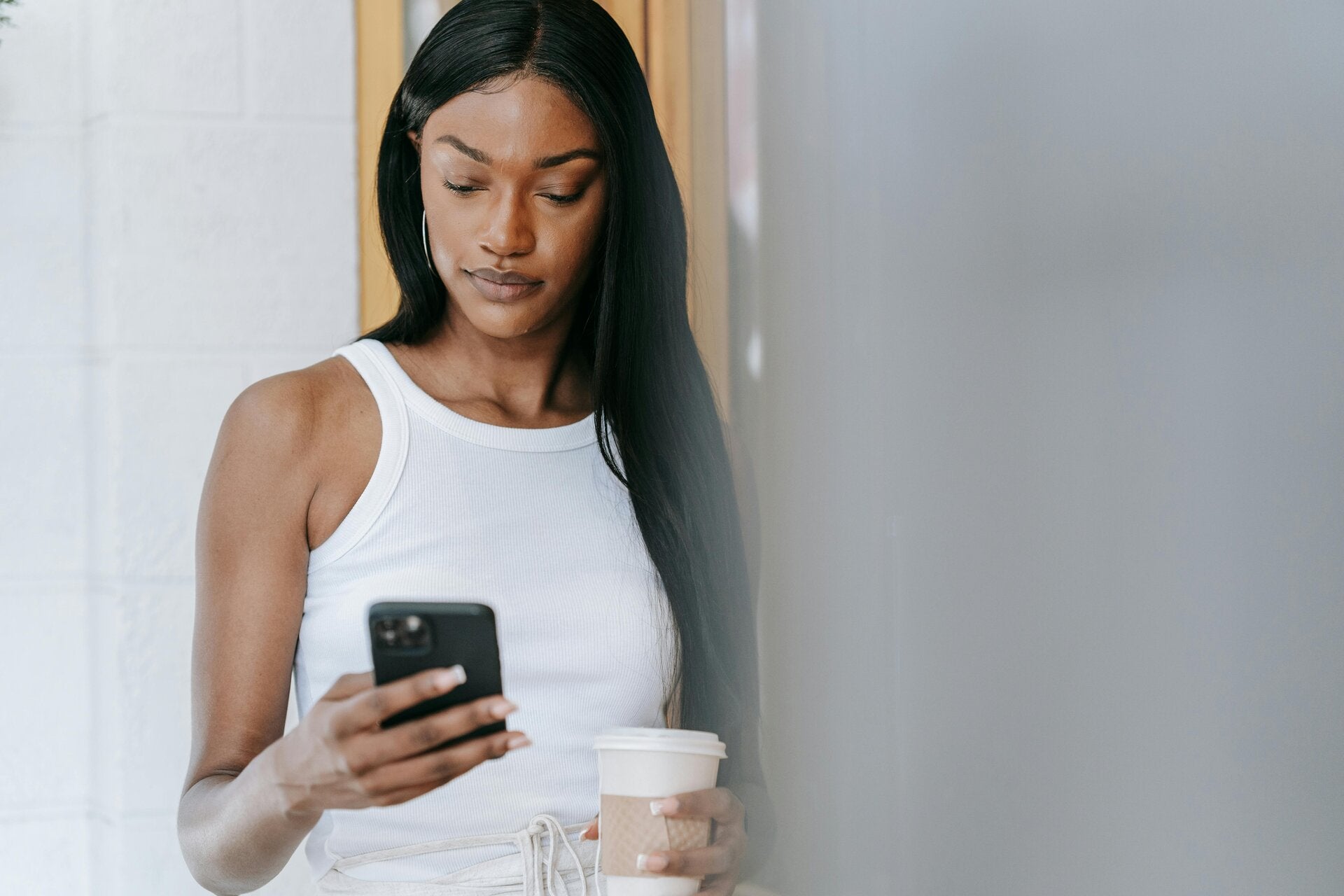 woman leaning against a wall holding a coffee cup looking at smartphone