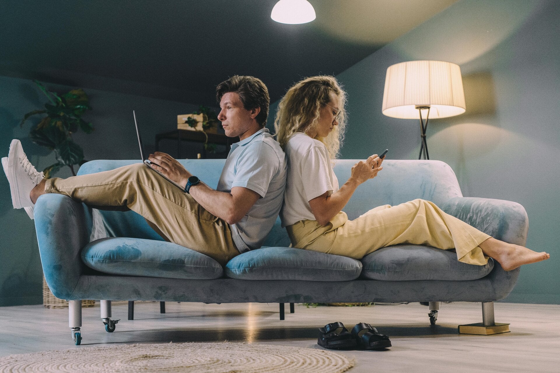 a couple sitting on a sofa with their backs against each other. one is holding a laptop and the other is holding a smartphone