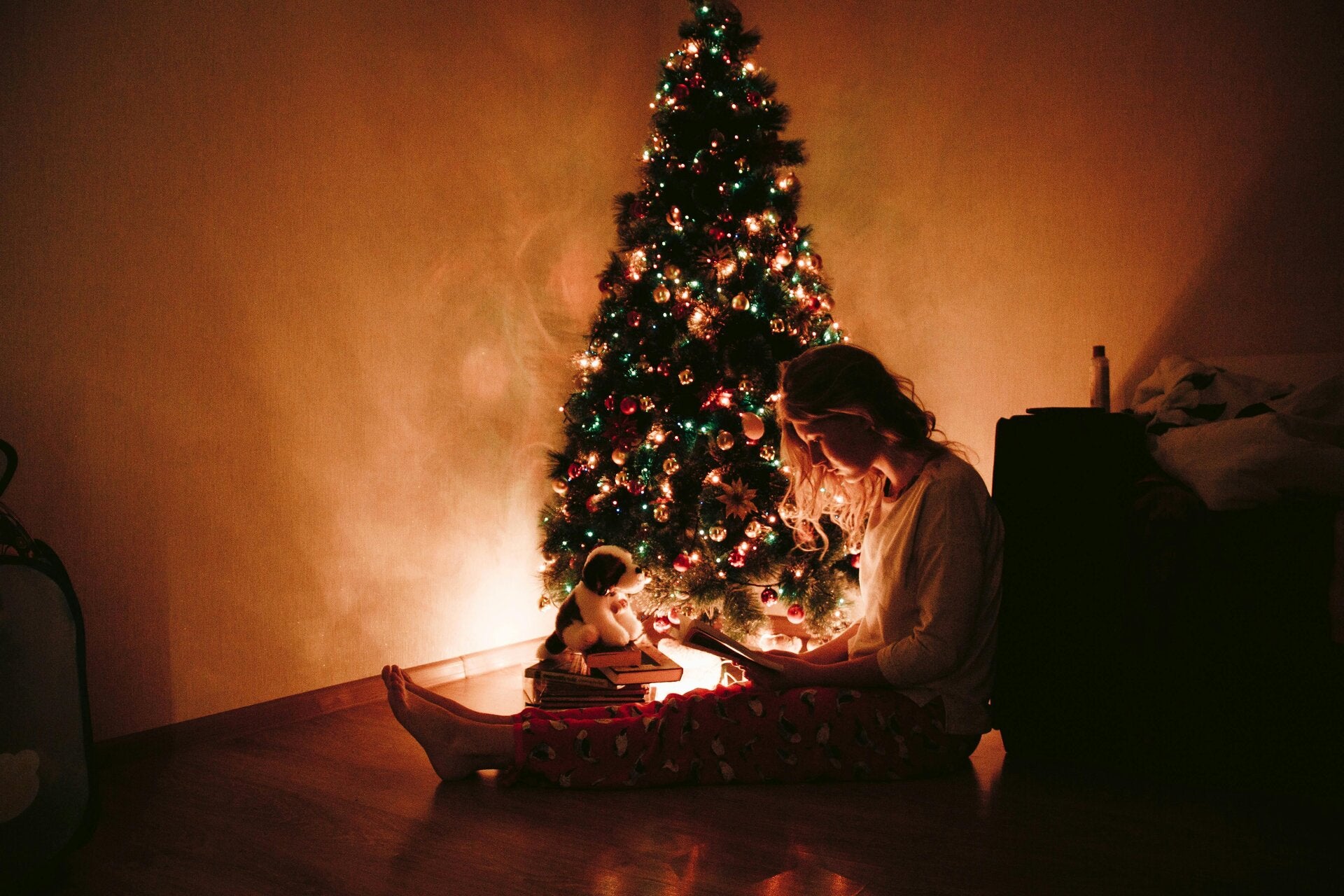 An image of a yonger women in the corner of a room sitting on the floor next to a Christmas tree and scrolling on her phone.The only light  in the room comes for the lights on the tree.