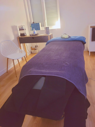 On y voit la pièce de massage avec la table de soin désolée d'un plaid bleu canard et d'un lotus. Une console avec les accessoires de massage et une chaise blanche.