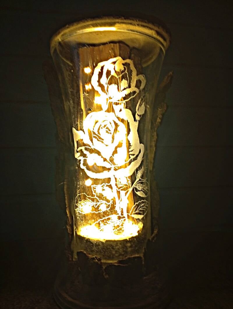 Handgefertigtes Lichtobjekt mit gravierter Rosenvase, Naturholz, Baumrinde, Beton und warmweißer LED-Beleuchtung.