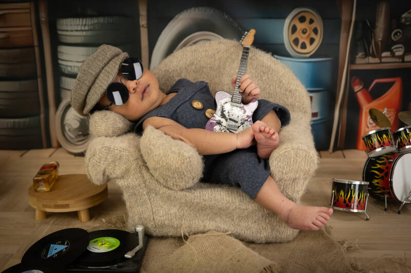 een lief klein baby jongen van 8 daagjes oud met een stoere zonnebril en een gitaar in zijn handjes met ernaast een drumstel. 