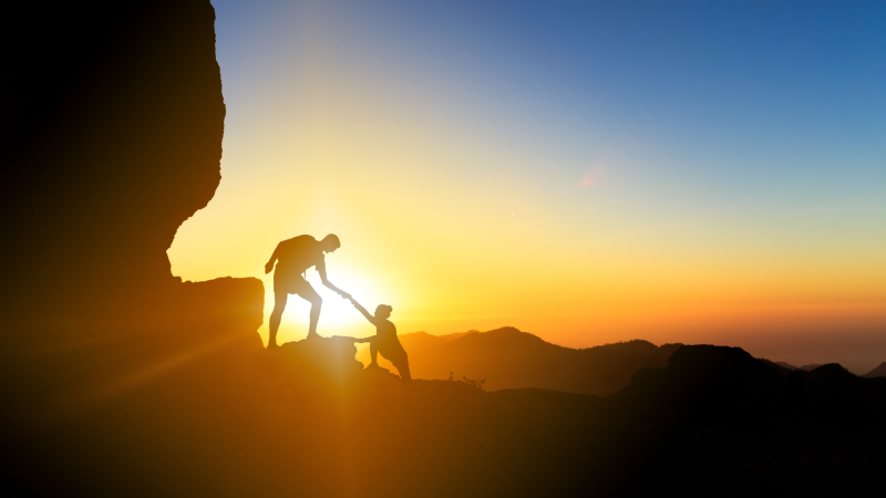 Foto van een man die een vrouw de berg op helpt bij opkomende zon. 