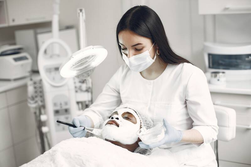 Cosmetóloga realizando una limpieza facial con mascarilla en una paciente con acné