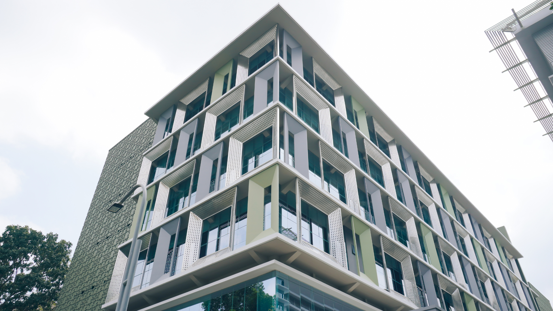 Low-angle view of a multi-story building with teal-and-white window bands, concrete facade, and a dark sloped roof under an overcast sky