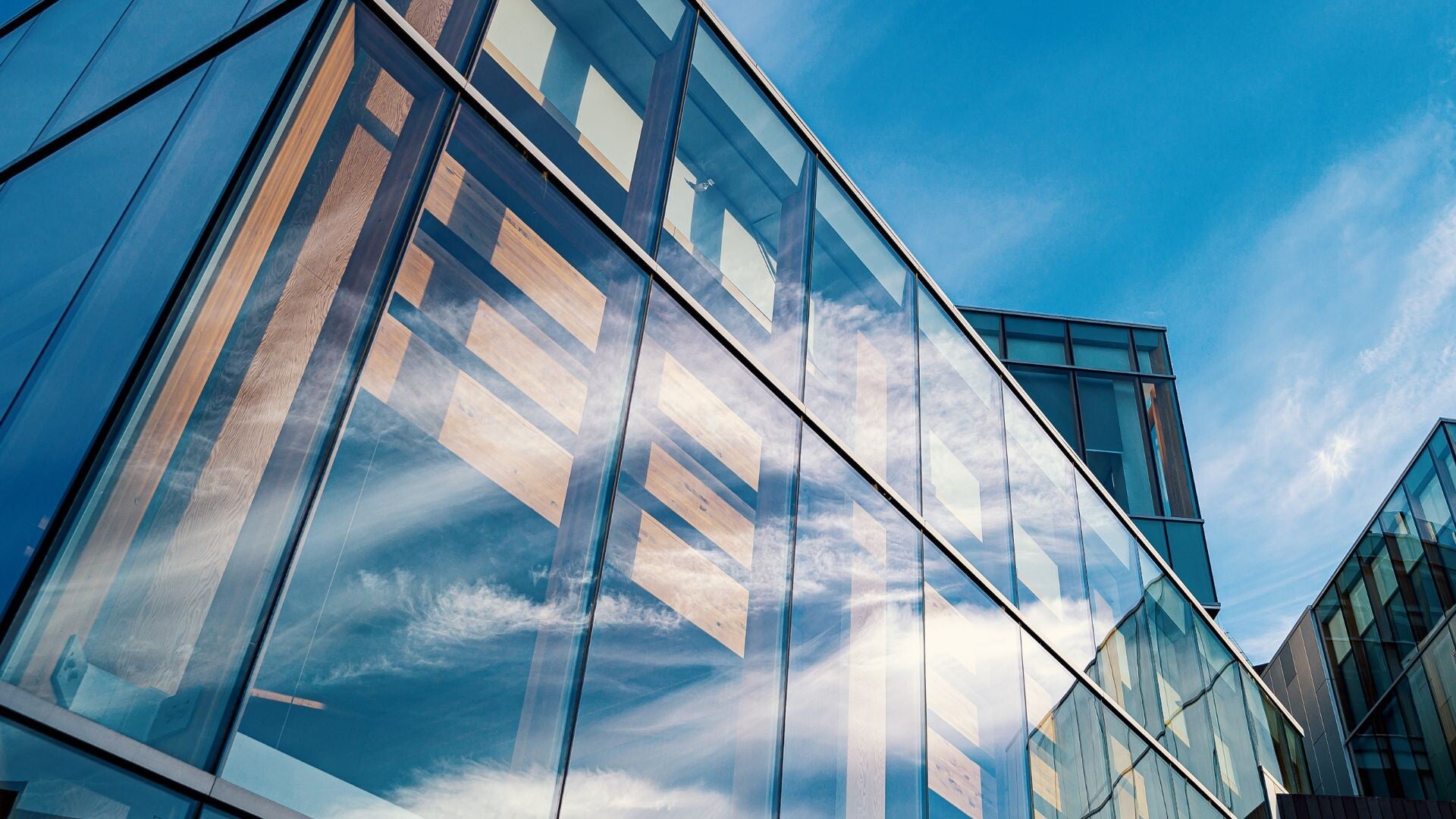 Low-angle view of a modern building with glass panels and vertical metal frames, reflecting clouds and sky, with other high-rise structures in the background.