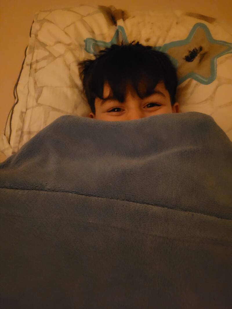 een jongen ligt in bed en zijn lachend gezichtje zit half onder een deken verstopt