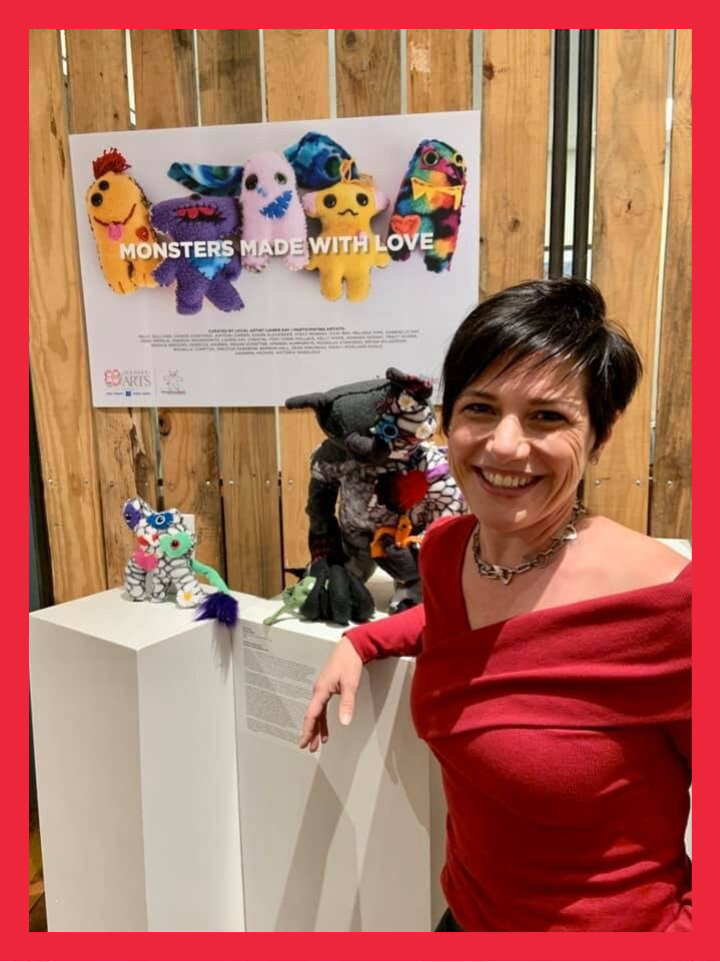 Laurie smiling beside a display of colorful handmade monsters and artwork.