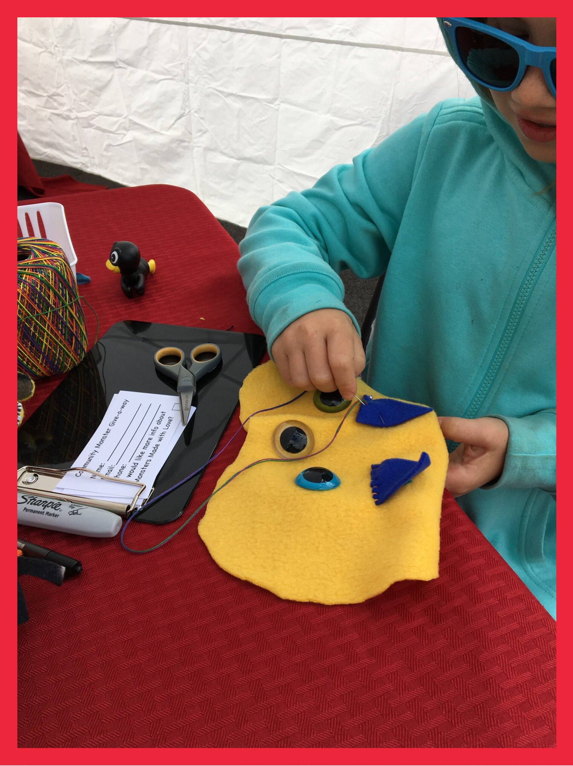 A child’s hand stitching a yellow handmade monster with blue felt details, button eyes, and a red tongue at a craft table.