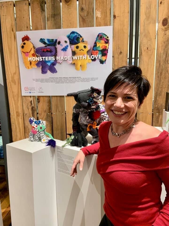 Laurie smiling in a red shirt beside a display of colorful handmade monsters and artwork.