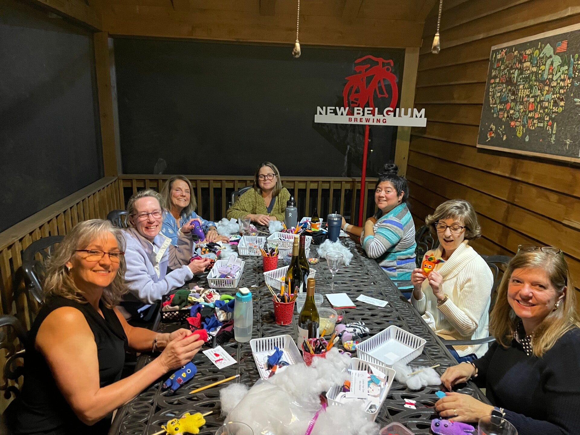 Eight adults seated around a table with colorful handmade plush monsters, fabric, stuffing, and craft supplies.