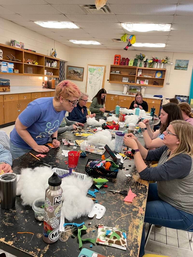 People gathered around a table with fabric, stuffing, handmade plush creations, and craft supplies.