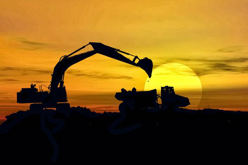 Silhouette of construction equipment, including an excavator and a dump truck, working against the backdrop of a setting sun.