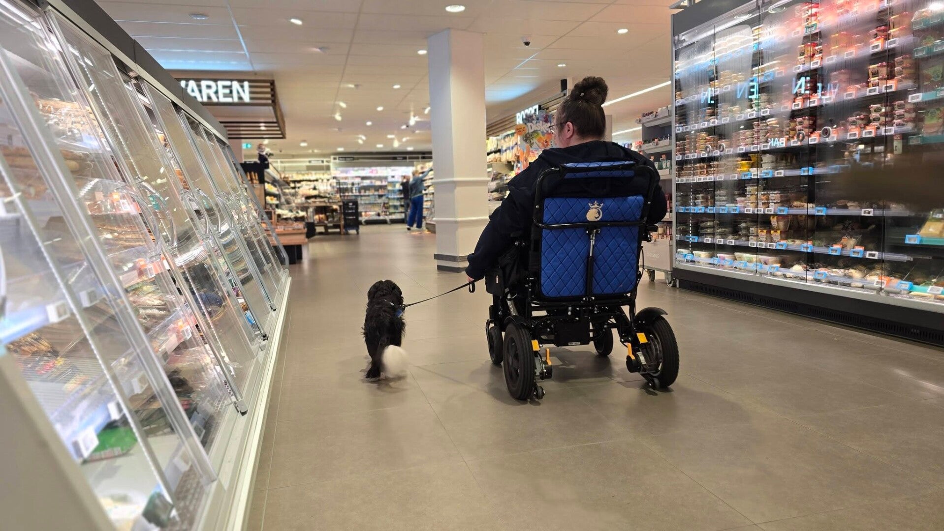 In de winkel, iemand in een rolstoel waar een hulphond naast loopt.