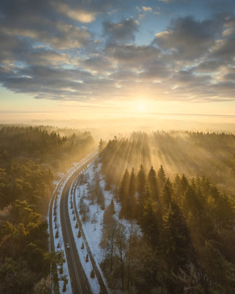 Beautiful aerial fine-art photo of a curving road throuh a winter forest with sunrays breaking through the fog