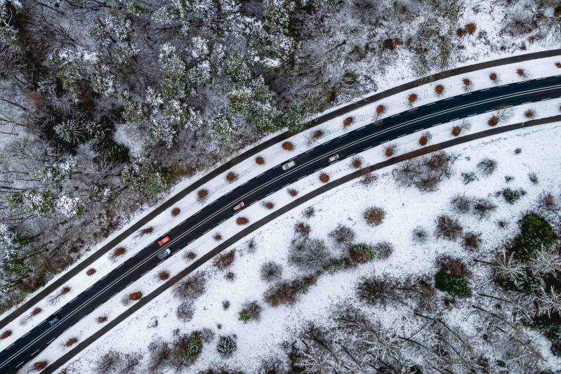 Top down aerial photograph of a black road through a snowy forest with 6 cars on the road.