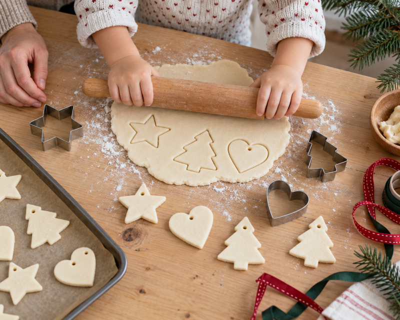 Kleines Kind rollt Salzteig aus und sticht Sterne, Herzen und Tannenbäume als Weihnachtsanhänger aus.