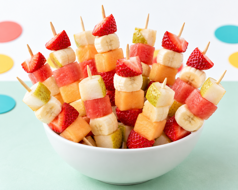 Bunte Obstspieße mit Erdbeeren, Banane, Birne, Wassermelone und Melone in einer weißen Schale für einen Kindergeburtstag.