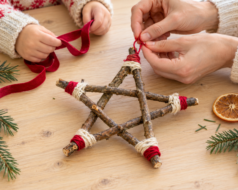 Erwachsene und Kind binden einen Weihnachtsstern aus kleinen Zweigen mit roter und naturfarbener Kordel zusammen