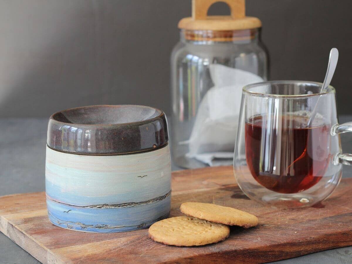 Irish stoneware tea bag holder with hand-carved coastal landscape and tiny birds in blue and light blue glaze, next to a cup of tea