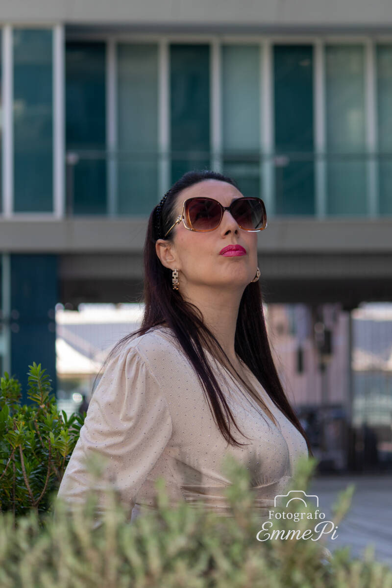 Donna con occhiali da sole posa all’aperto davanti a un edificio moderno, ritratto urbano elegante con luce naturale e sfondo architettonico sfocato.