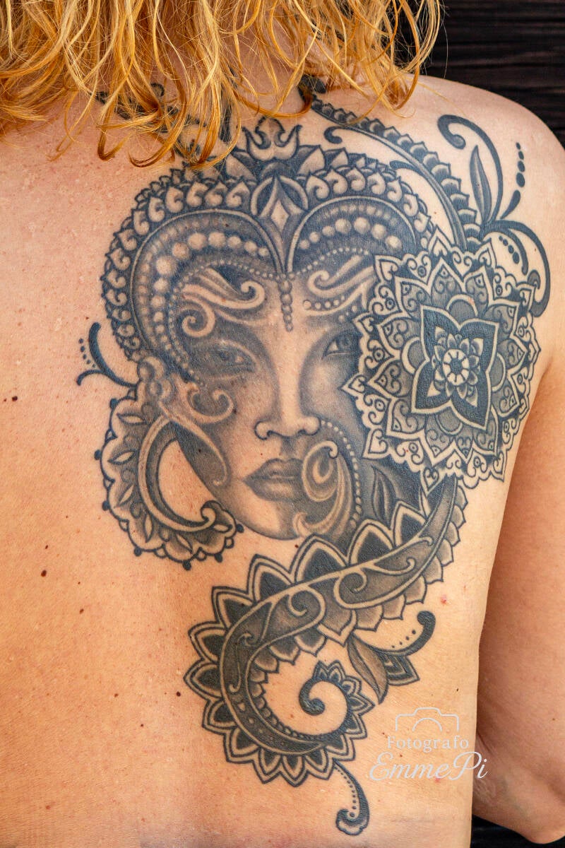 Dettaglio di un grande tatuaggio ornamentale sulla schiena raffigurante un volto femminile intrecciato a motivi floreali e mandala.