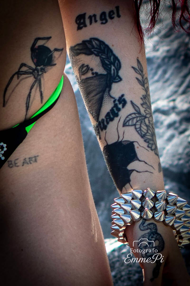 Dettaglio di braccio tatuato con scritte, ragno e bracciale borchiato in stile punk.
