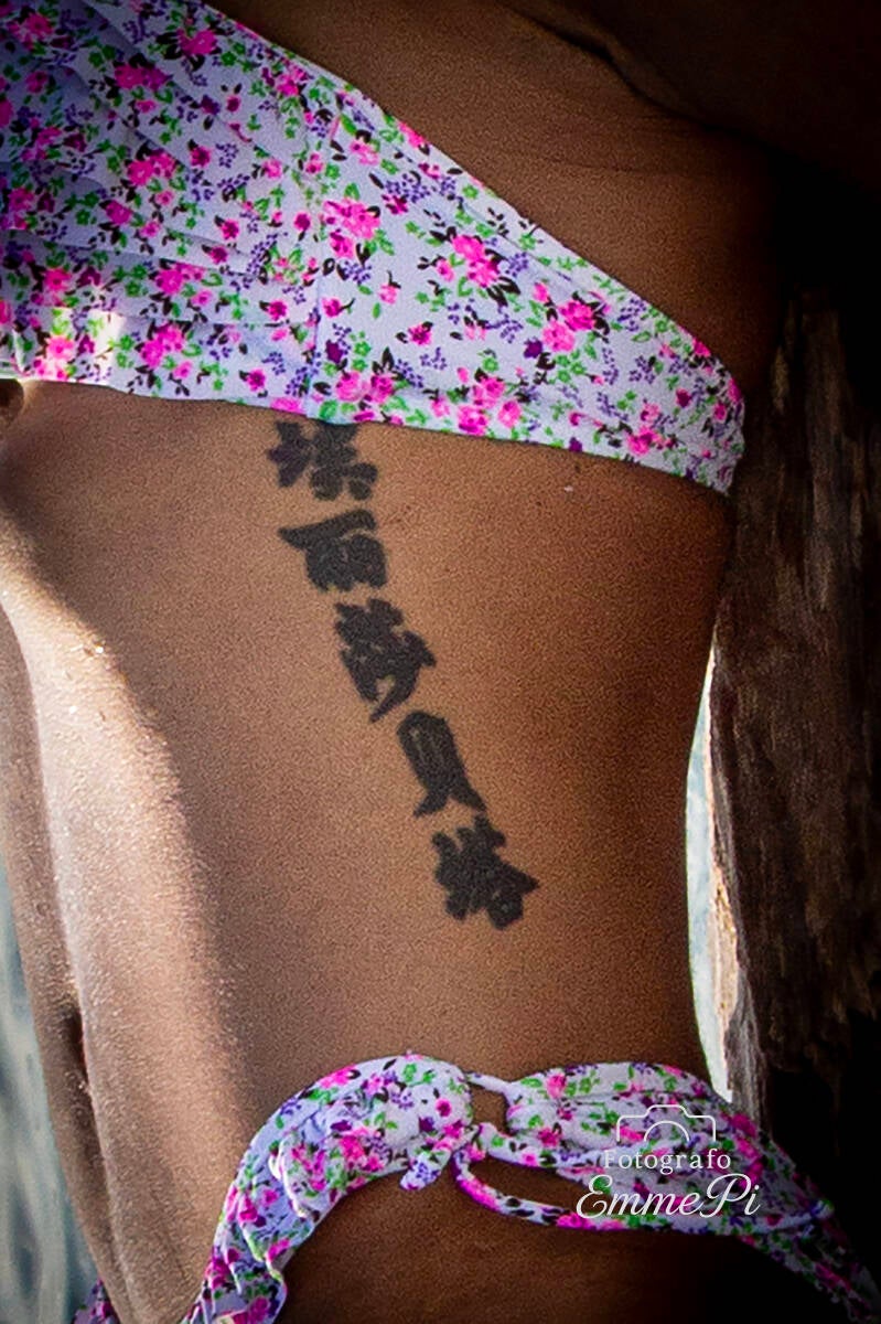 Dettaglio del fianco di una modella in bikini floreale con tatuaggio verticale in caratteri orientali sul costato.