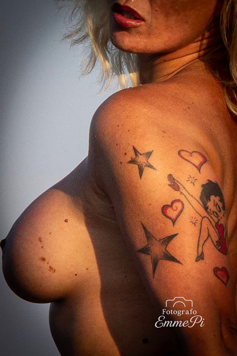Ritratto parziale di modella tatuata con disegno di Betty Boop, cuori e stelle colorate sulla spalla e sul petto.