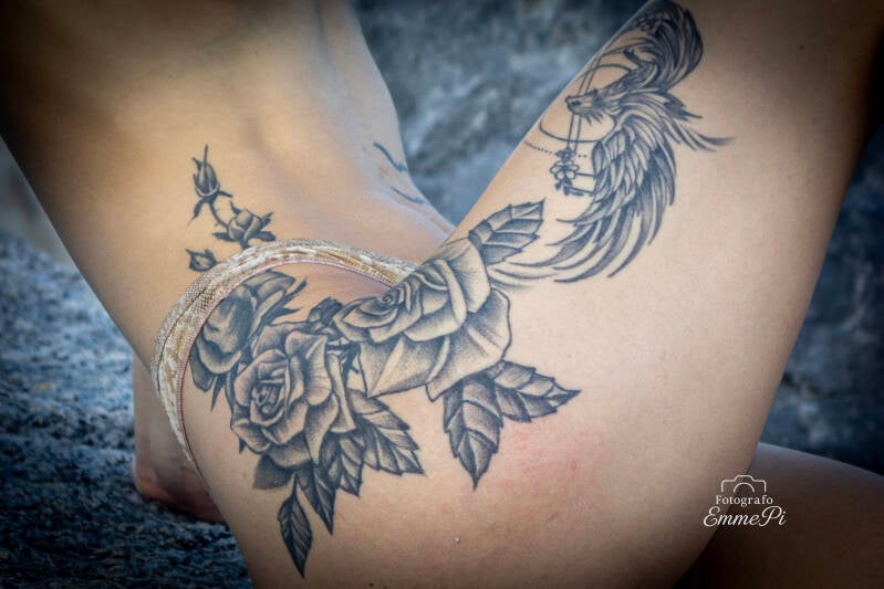 Tatuaggio con rose e fenice stilizzata sul fianco femminile fotografato in luce naturale sugli scogli.