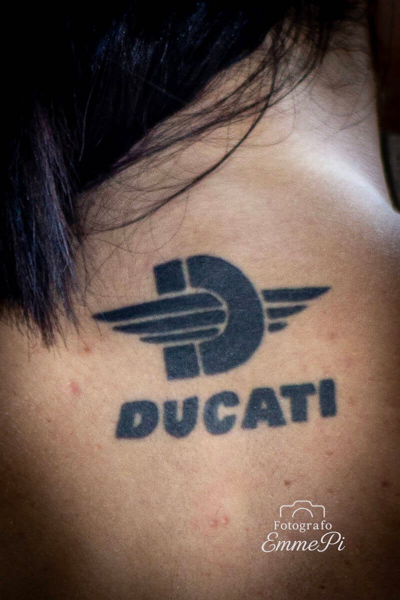 Dettaglio di un tatuaggio Ducati nero sulla schiena di una modella dai capelli scuri.