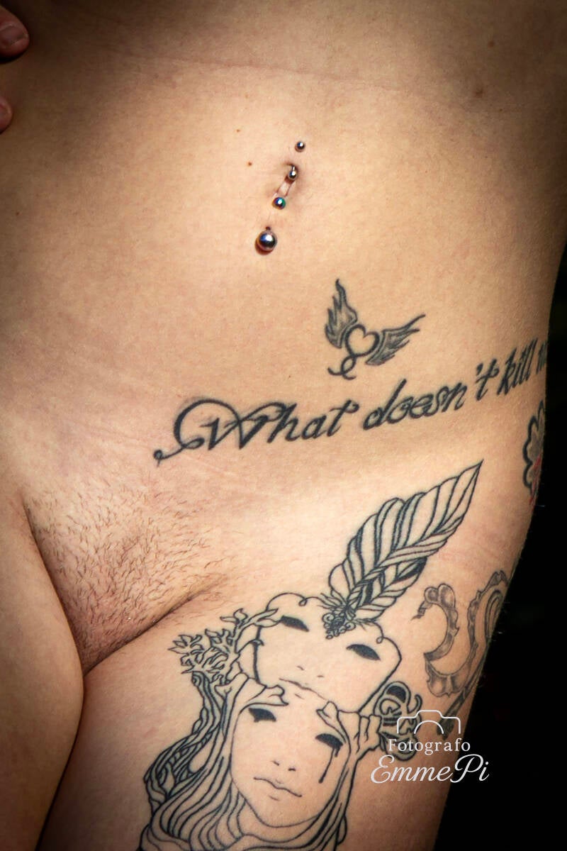 Dettaglio di tatuaggi su addome femminile con scritta calligrafica, piercing all’ombelico e figura illustrata sulla coscia.