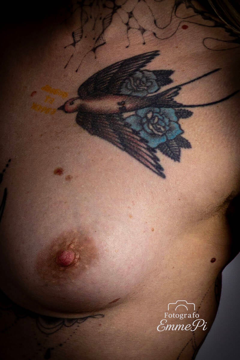 Primo piano di tatuaggio a forma di rondine con rose blu sul petto femminile illuminato da luce contrastata.