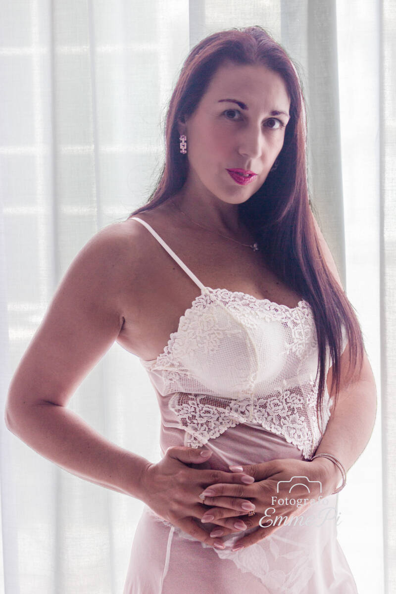 Ritratto boudoir di donna in lingerie bianca, sguardo in camera con mani sul ventre