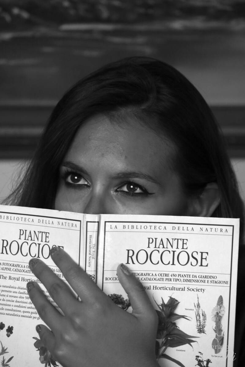 Ritratto in bianco e nero di Dafne che osserva l'obiettivo con sguardo malizioso da dietro un libro aperto.